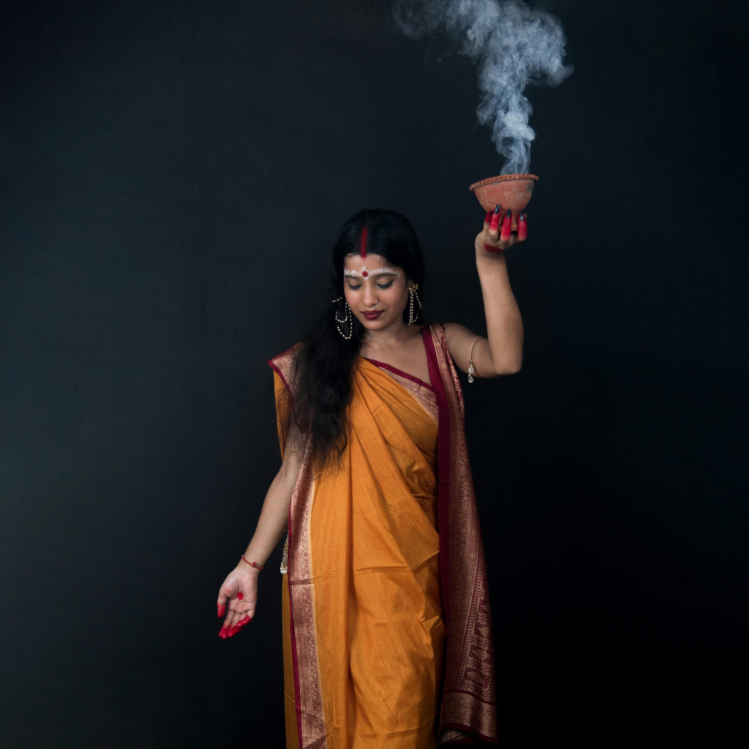 A woman in traditional attire holding a pot with smoke in a studio setting against a black background.