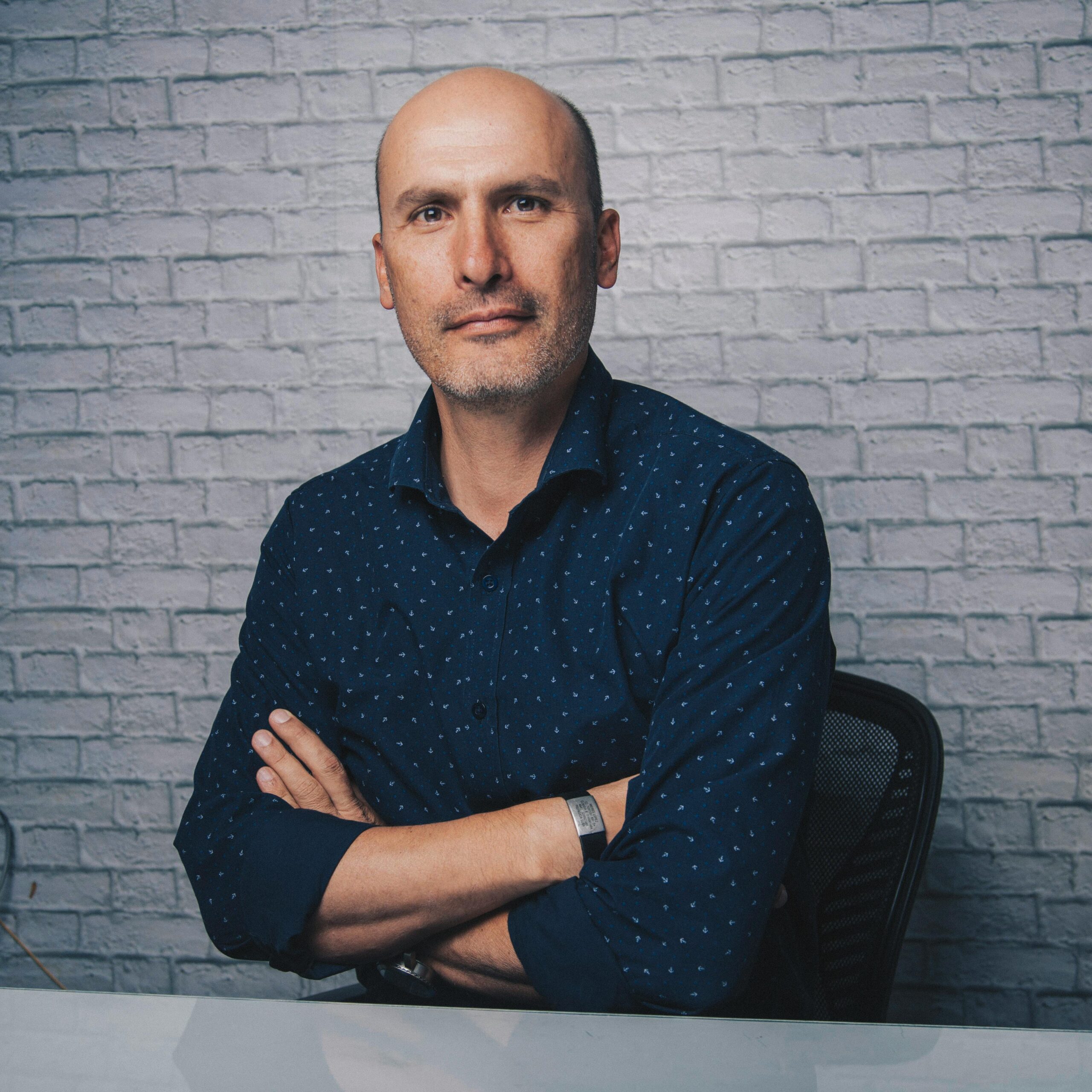 Positive bald male in blue shirt sitting at shiny desk with crossed arms while looking at camera in white room